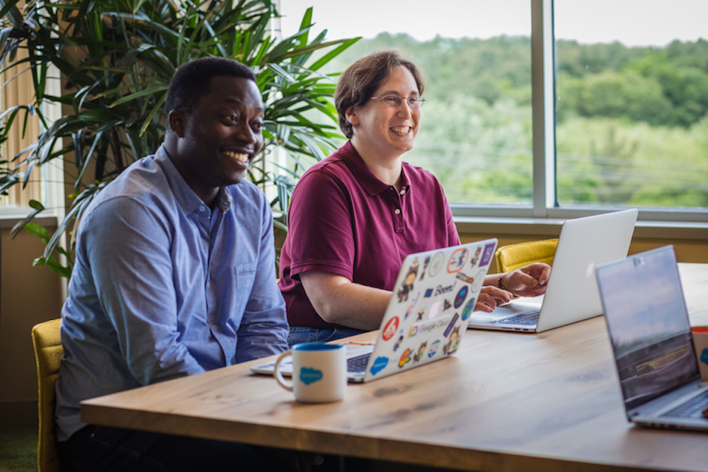 A smiling man sits in front of a laptop computer covered with stickers, next to a smiling woman who is also sitting in front of a laptop.