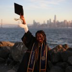 A woman with dark braided hair wearing a graduation gown stands in front of a body of water with a city skyline visible in the background, holding her mortarboard up in celebration.